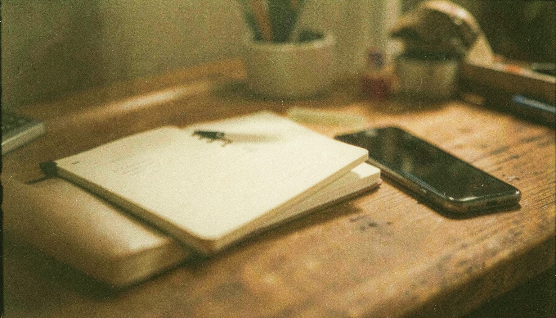 A warm, softly lit desk scene with a notebook, pen, and a smartphone resting on a wooden surface, shot on grainy analog film with muted earth tones and gentle shadows.