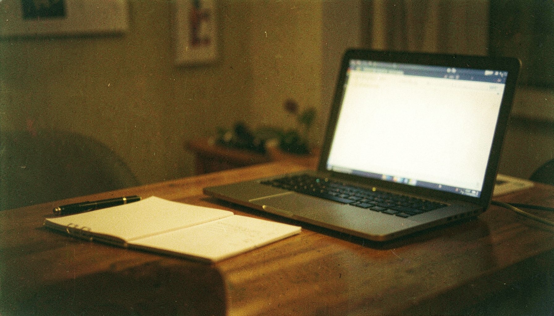 A warm-toned analog film photograph of a wooden desk with a notebook, pen, and a laptop screen glowing softly in a dim room, with visible film grain and a cozy, focused atmosphere.