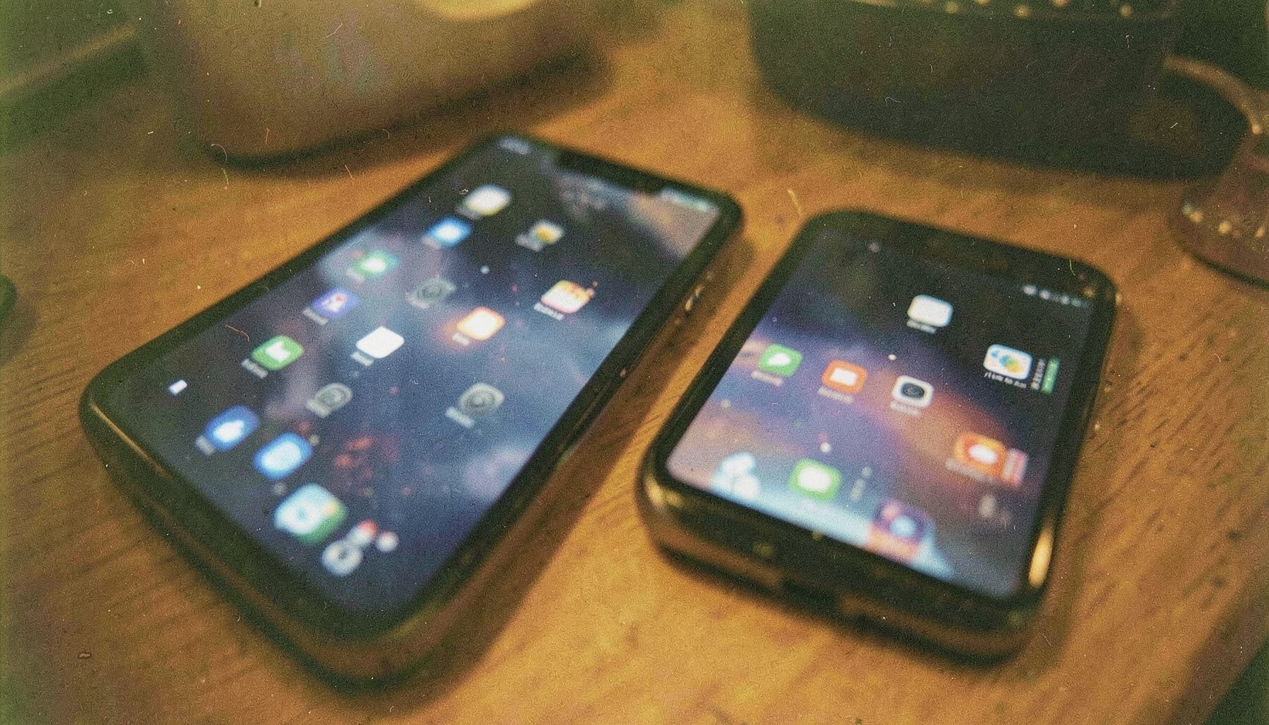 A close-up analog film photograph of a tablet and smartphone resting on a wooden desk, both screens glowing softly with app icons visible. Warm amber and deep navy tones dominate, with visible film grain and a cozy, lived-in atmosphere.