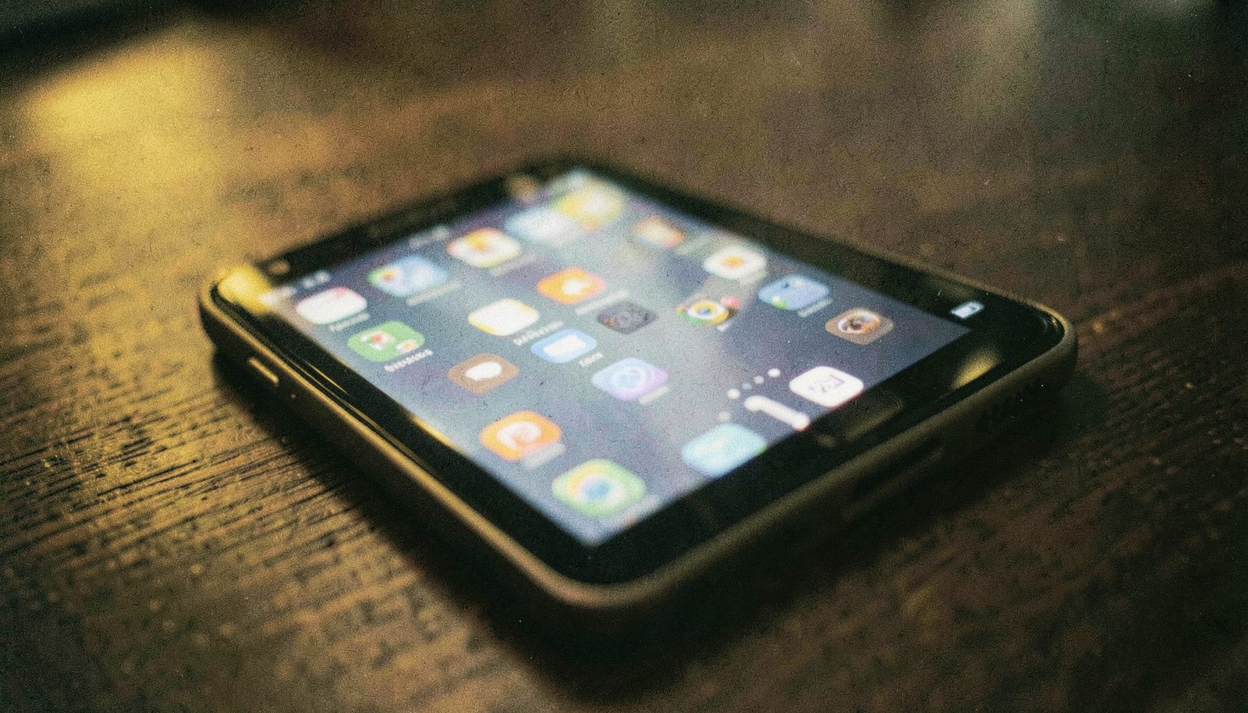 A close-up analog film photograph of a modern Android smartphone resting on a dark wooden table, its screen glowing softly with colorful app icons. Warm amber tones blend with deep charcoal shadows, and visible film grain adds a tactile, editorial feel.