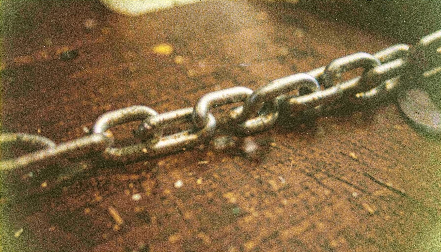 A grainy analog film close-up of interlinked metal chain links on a dark wooden desk, warm brown and steel gray tones.